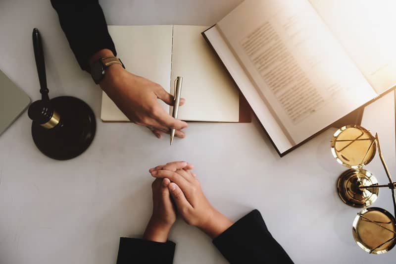 Find-an-Attorney-4 Hands on a white table with gavel, notebook, textbook, and brass scale